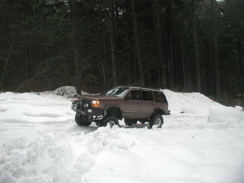 1985 Ford Bronco - A lifted, brown SUV with off-road tires is driving through deep snow in a forest setting. The vehicle has a custom bumper and appears to be…