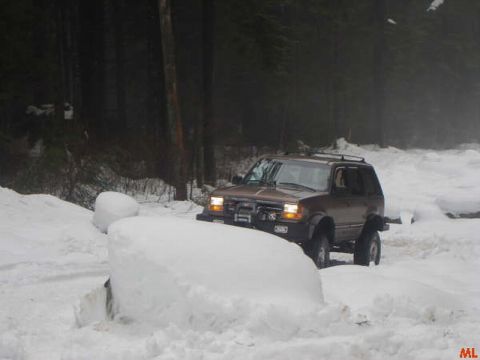 1985 Ford Bronco - A brown SUV drives through deep snow on a wooded road, its headlights on. The vehicle appears to be navigating a challenging, snow-covered…