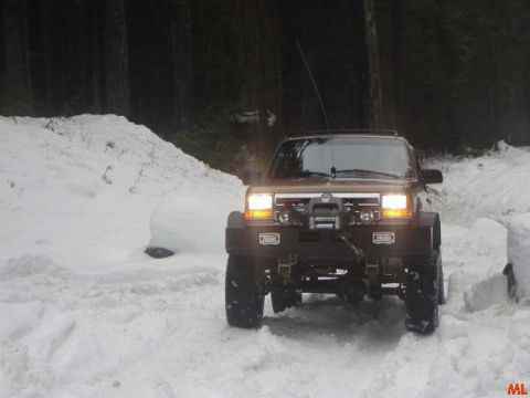 1985 Ford Bronco - A lifted, brown SUV with a winch on the front bumper is driving through deep snow in a wooded area. The vehicle's headlights are on, illumi…