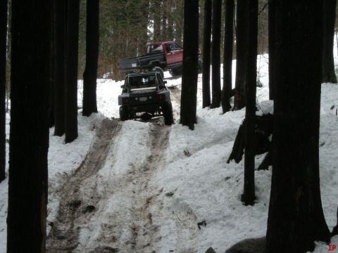 1985 Ford Bronco - Two off-road vehicles, a red pickup truck and a black Jeep-style SUV, navigate a snowy, wooded trail. The Jeep is in the foreground, drivin…