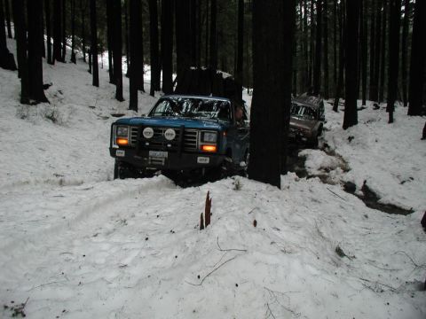 1985 Ford Bronco - Two off-road vehicles navigate a snow-covered forest trail. The lead blue SUV is equipped with auxiliary lights and a brush guard.