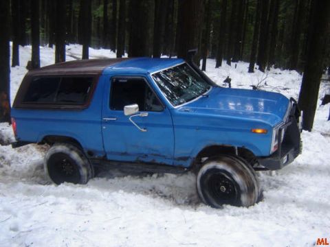 1985 Ford Bronco - A blue Ford Bronco with a brown top is stuck in the snow in a forest. The vehicle's front tire is partially submerged in snow, and the wind…