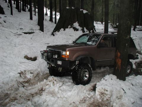 1985 Ford Bronco - A lifted, brown SUV with large off-road tires is navigating through a snowy forest, its front bumper equipped with a winch and auxiliary li…