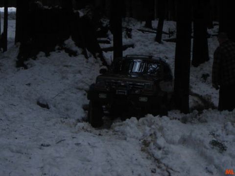 1985 Ford Bronco - A dark-colored SUV is partially submerged in snow in a wooded area. A person in a plaid shirt stands to the right of the vehicle.