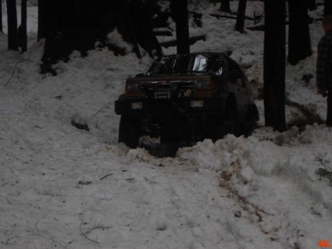 1985 Ford Bronco - A dark-colored SUV is stuck in deep snow on a wooded trail. The vehicle appears to be a Ford Bronco, with its front end partially buried in…
