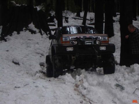 1985 Ford Bronco - A lifted, modified SUV with a winch is driving through deep snow in a wooded area. A person in a plaid shirt stands to the right of the veh…