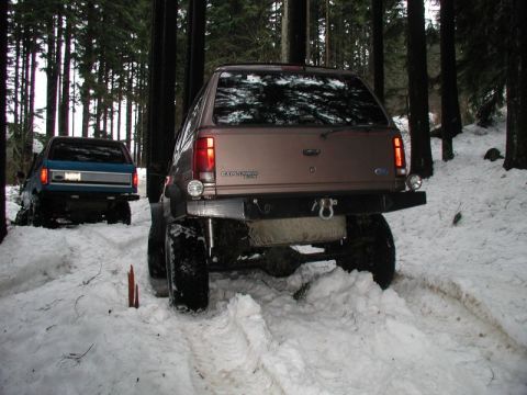 1985 Ford Bronco - Two modified SUVs drive through a snowy forest. The vehicle in the foreground is a brown SUV with large tires and a lifted suspension. A bl…