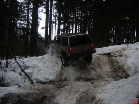 1985 Ford Bronco - A dark colored SUV is driving through deep snow and slush in a wooded area. Water and snow are splashing up from the tires as it navigates…