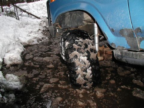 1985 Ford Bronco - A blue SUV's front tire is stuck in muddy, icy water on a snowy trail. The vehicle's fender and side are visible, showing some rust and wea…