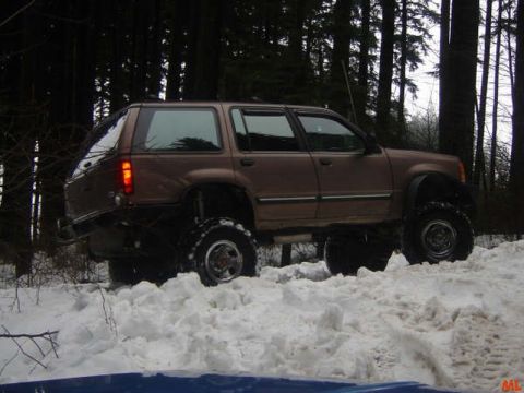 1985 Ford Bronco - A lifted brown SUV is driving through deep snow in a forest. The vehicle has large off-road tires and appears to be a Ford Explorer.