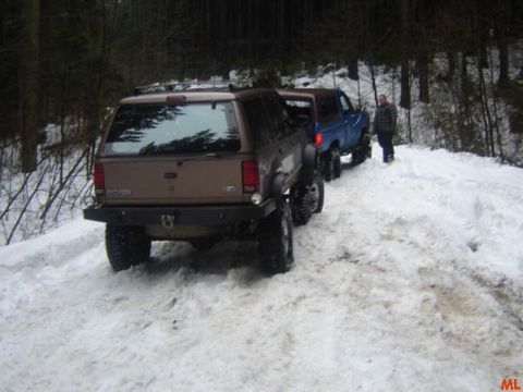 1985 Ford Bronco - Two off-road vehicles are parked on a snow-covered forest road. The lead vehicle is a tan SUV, and behind it is a blue SUV with a person st…