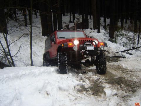 1985 Ford Bronco - A red Jeep Wrangler is driving through a snowy forest road, with its headlights on and tires deep in the snow.