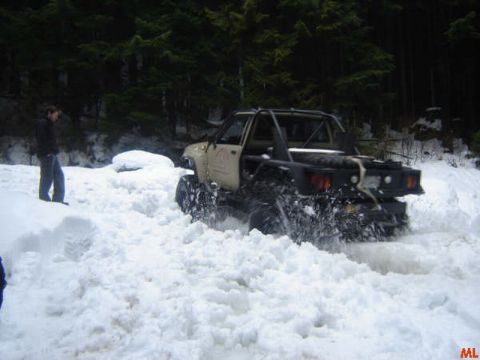 1985 Ford Bronco - A tan off-road truck with a roll cage and a flatbed is driving through deep snow in a wooded area. A person stands to the left of the truck…