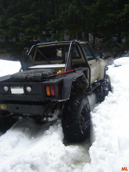 1985 Ford Bronco - A modified pickup truck with large tires is driving through deep snow in a wooded area. The truck has a flatbed with a spare tire mounted o…