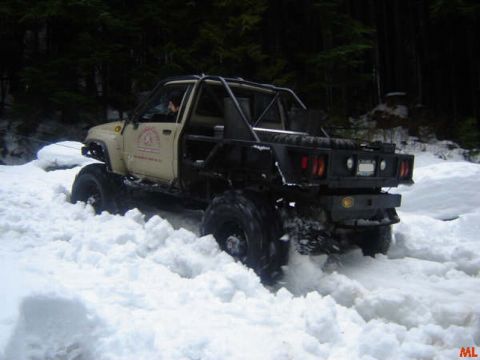 1985 Ford Bronco - A modified beige and black pickup truck with a flatbed is driving through deep snow in a wooded area. The truck has large tires and a roll…