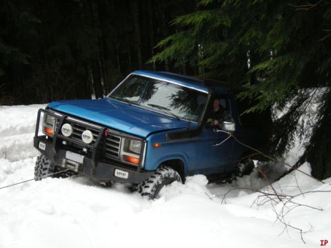 1985 Ford Bronco - A blue 1980s Ford pickup truck is stuck in deep snow in a wooded area, with a winch cable attached to the front bumper. The driver is visib…