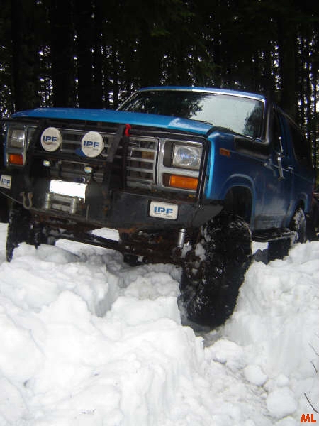 1985 Ford Bronco - A blue Ford Bronco is driving through deep snow in a wooded area. The front of the vehicle shows off-road modifications including IPF light…