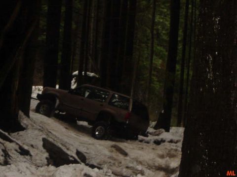 1985 Ford Bronco - A brown SUV drives on a snowy, wooded trail. The vehicle is angled uphill, with its front wheels turned slightly to the left.