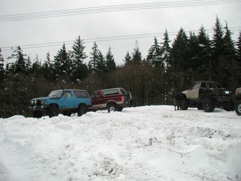 1985 Ford Bronco - A blue Ford Bronco is parked in the snow next to a maroon Ford Bronco, with a white custom pickup truck and another vehicle visible further…