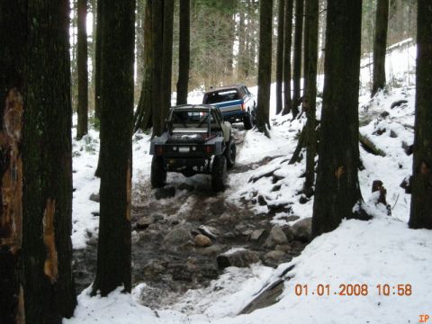 1985 Ford Bronco - Two off-road vehicles navigate a snow-covered, rocky trail through a dense forest. The lead vehicle is a modified, dark-colored buggy, foll…