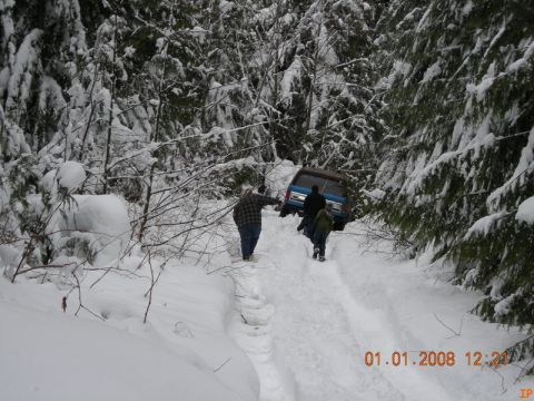 1985 Ford Bronco - Three people walk behind a blue SUV stuck in a snowy forest path. The vehicle appears to be a Ford Bronco.