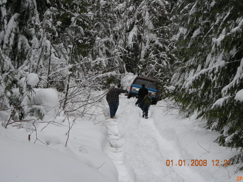 Blue SUV stuck in snow with three people walking behind it in a forest.