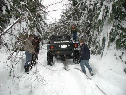 1985 Ford Bronco - A group of people are gathered around a lifted, flatbed truck stuck in a snowy forest trail. The truck appears to be winching itself out or…