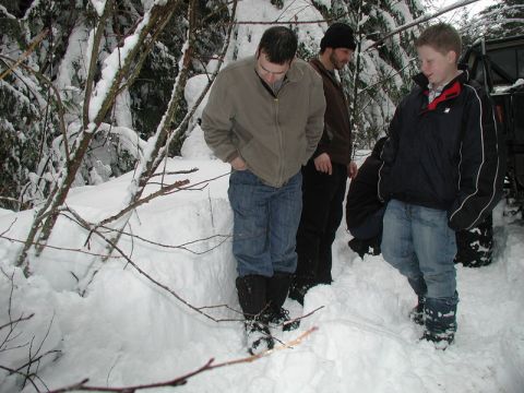 1985 Ford Bronco - Four people stand in a snowy forest, with trees and snow-covered branches surrounding them. A large pile of snow is in the foreground.
