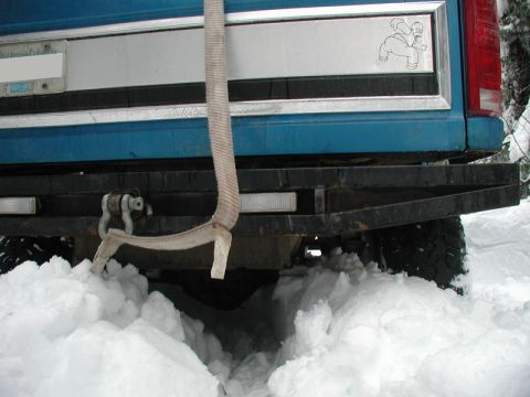 1985 Ford Bronco - The rear of a blue Ford truck is stuck in deep snow, with a tow strap attached to a shackle on the heavy-duty bumper. A sticker of Homer Si…