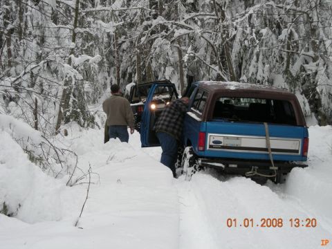 1985 Ford Bronco - Two blue Ford Broncos are stuck in deep snow in a wooded area. One Bronco has its rear facing the camera, showing a brown roof and a tow st…