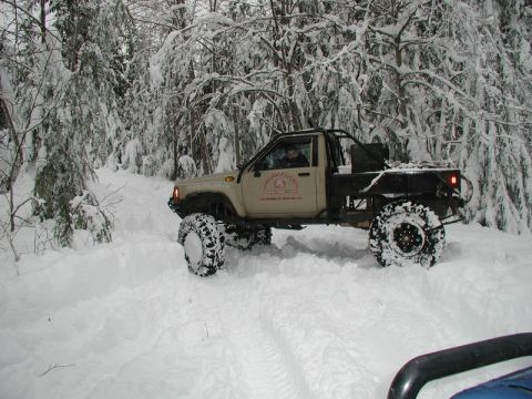1985 Ford Bronco - A lifted, tan pickup truck with large off-road tires navigates a snow-covered forest road. The truck has a custom flatbed and a roll cage v…