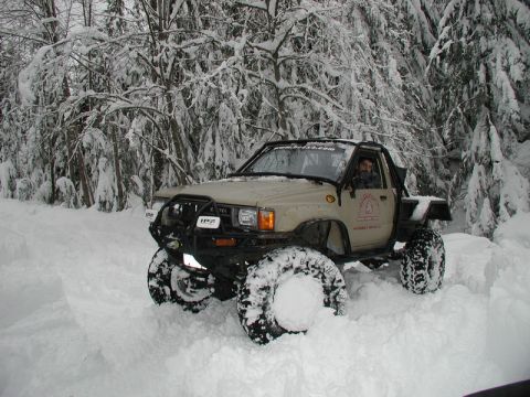 1985 Ford Bronco - A tan Toyota pickup truck with oversized tires navigates a snow-covered forest road. The truck features a custom front bumper with IPF ligh…