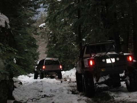1985 Ford Bronco - Two off-road vehicles drive through a snow-covered forest road. The vehicle in the foreground is a modified pickup truck with a roll cage a…