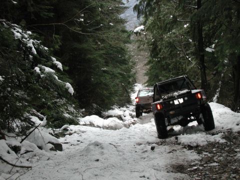 1985 Ford Bronco - Two off-road vehicles navigate a snow-covered trail through a dense forest. The lead vehicle is a dark-colored, modified Jeep-style SUV, wh…