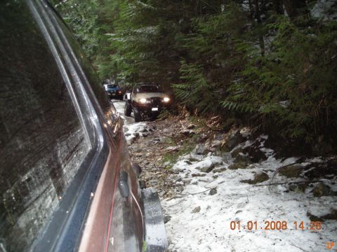 1985 Ford Bronco - A line of off-road vehicles navigates a snowy, wooded trail. The lead vehicle is a white SUV with its headlights on, followed by a blue SUV…