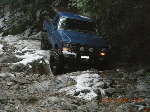 1985 Ford Bronco - A blue Ford Bronco is navigating a rocky, snow-covered trail in a wooded area. The vehicle has large tires and auxiliary lights mounted on…
