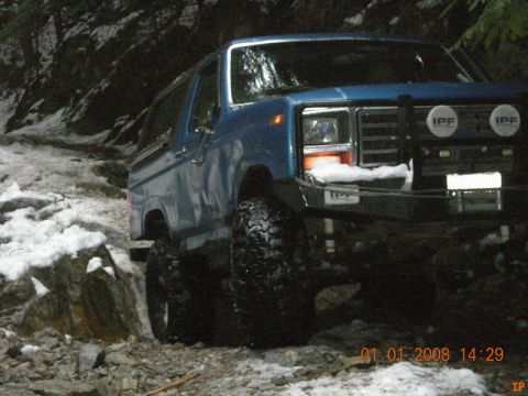 1985 Ford Bronco - A blue Ford Bronco is parked on a snowy, rocky trail in a wooded area. The vehicle has large off-road tires and IPF lights mounted on the f…