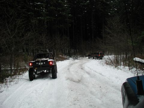 1985 Ford Bronco - Two off-road vehicles navigate a snow-covered forest road. The vehicle in the foreground is a modified flatbed truck with prominent red tai…