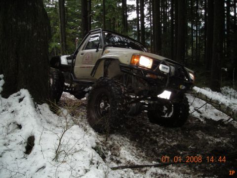 1985 Ford Bronco - A lifted off-road vehicle with large tires navigates a snowy and muddy forest trail. The vehicle has a custom front bumper with lights and…