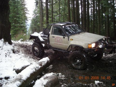 1985 Ford Bronco - A modified tan Toyota pickup truck with large off-road tires navigates a muddy and snowy forest trail. The truck has a roll cage and custom…