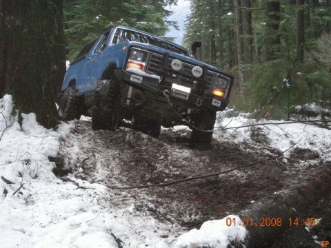 1985 Ford Bronco - A blue Ford Bronco is navigating a muddy, snow-covered trail in a dense forest. The vehicle is angled uphill, with its front tires deep in…