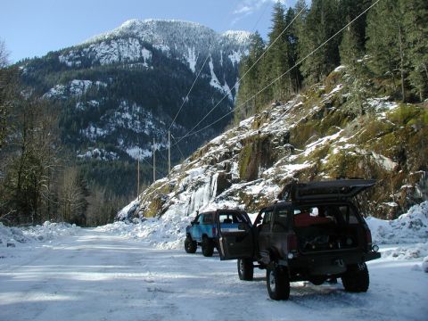 1985 Ford Bronco - Two off-road SUVs are parked on a snow-covered road in a mountainous, forested area. The SUV in the foreground has its rear hatch and drive…