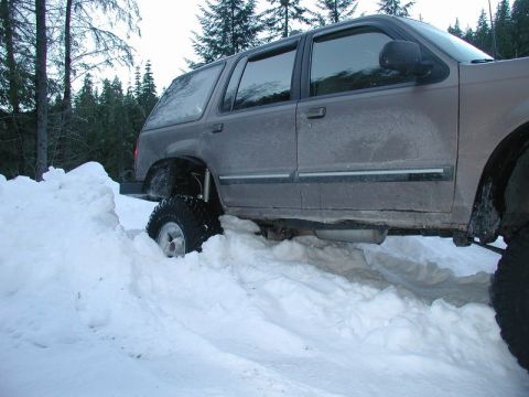 1985 Ford Bronco - A lifted, dark-colored SUV is stuck in deep snow with its front tire partially buried. The vehicle is surrounded by a snowy landscape with…