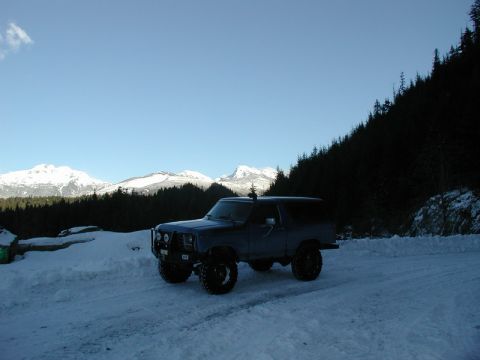 1985 Ford Bronco - A dark blue Ford Bronco is parked on a snow-covered road with a mountain range and forest in the background.