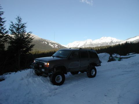 1985 Ford Bronco - A lifted, dark-colored SUV is parked on a snowy path in a mountainous, forested area with snow-capped peaks in the background.