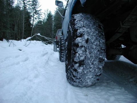 1985 Ford Bronco - A close-up, low-angle shot of the front tire of a blue Ford Bronco covered in snow, parked on a snowy path in a forest. Another vehicle is…