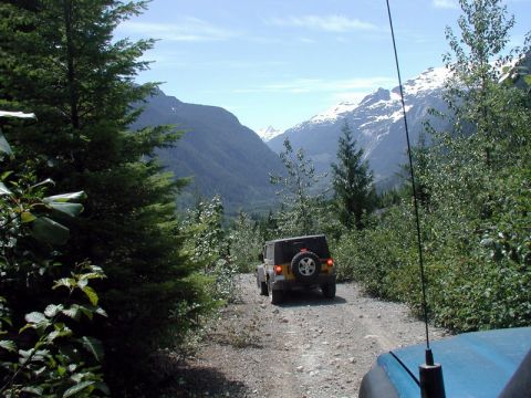1985 Ford Bronco - A yellow Jeep Wrangler drives down a gravel road in a mountainous, forested area with snow-capped peaks in the distance. The view is from a…