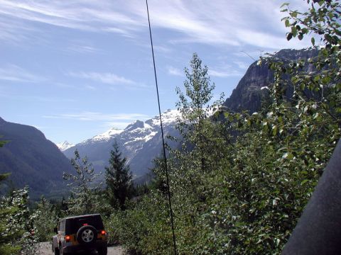 1985 Ford Bronco - A yellow Jeep Wrangler is driving on a gravel road through a mountain valley. The vehicle is heading away from the camera, with snow-capped…