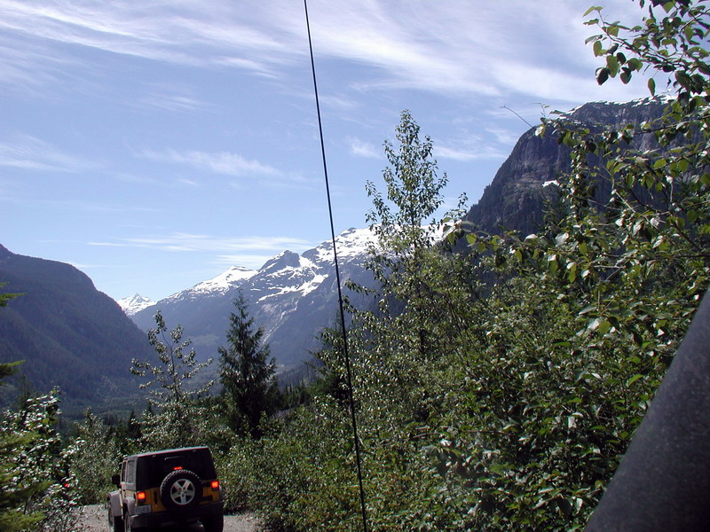 Yellow Jeep Wrangler driving on a mountain road with snow-capped peaks in the background.