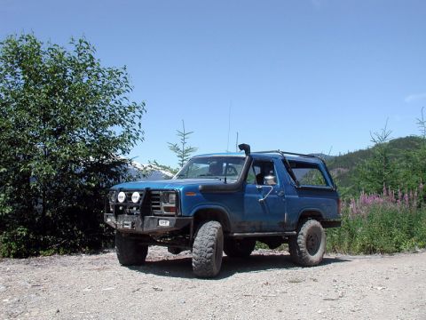 1985 Ford Bronco - A blue 1980s Ford Bronco with off-road modifications is parked on a gravel road in a wooded, mountainous area.
